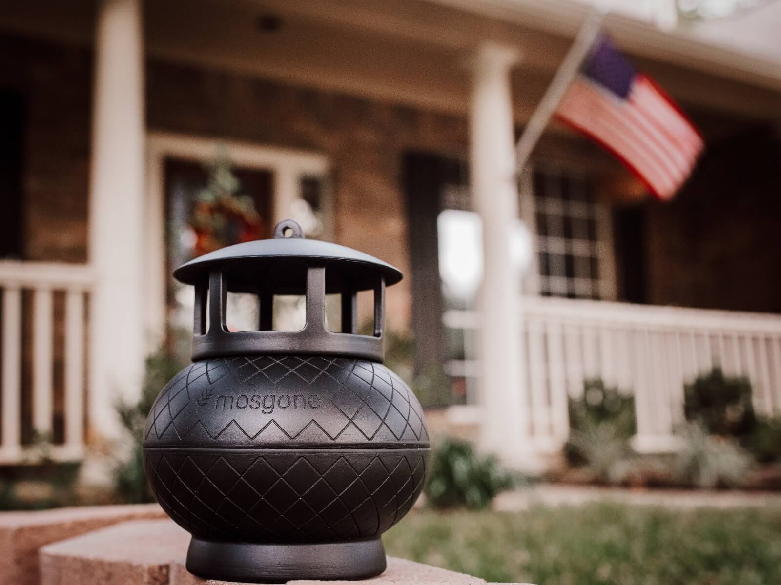 A black mosquito repellent lantern labeled mosgone sits on a porch ledge, with a house and an American flag in the blurred background.