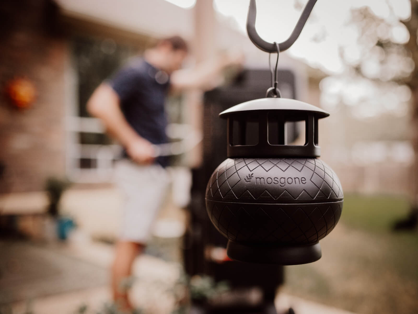 A black Mosgone mosquito repellent device hangs in focus outdoors, while a blurred man stands near a barbecue grill in the background.