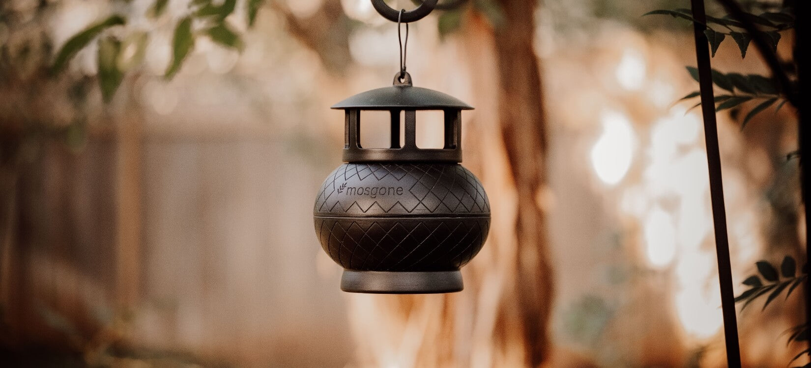 A black, lantern-shaped mosquito trap hangs outdoors, surrounded by blurred greenery and sunlight in the background. The word mosgone is visible on the front.