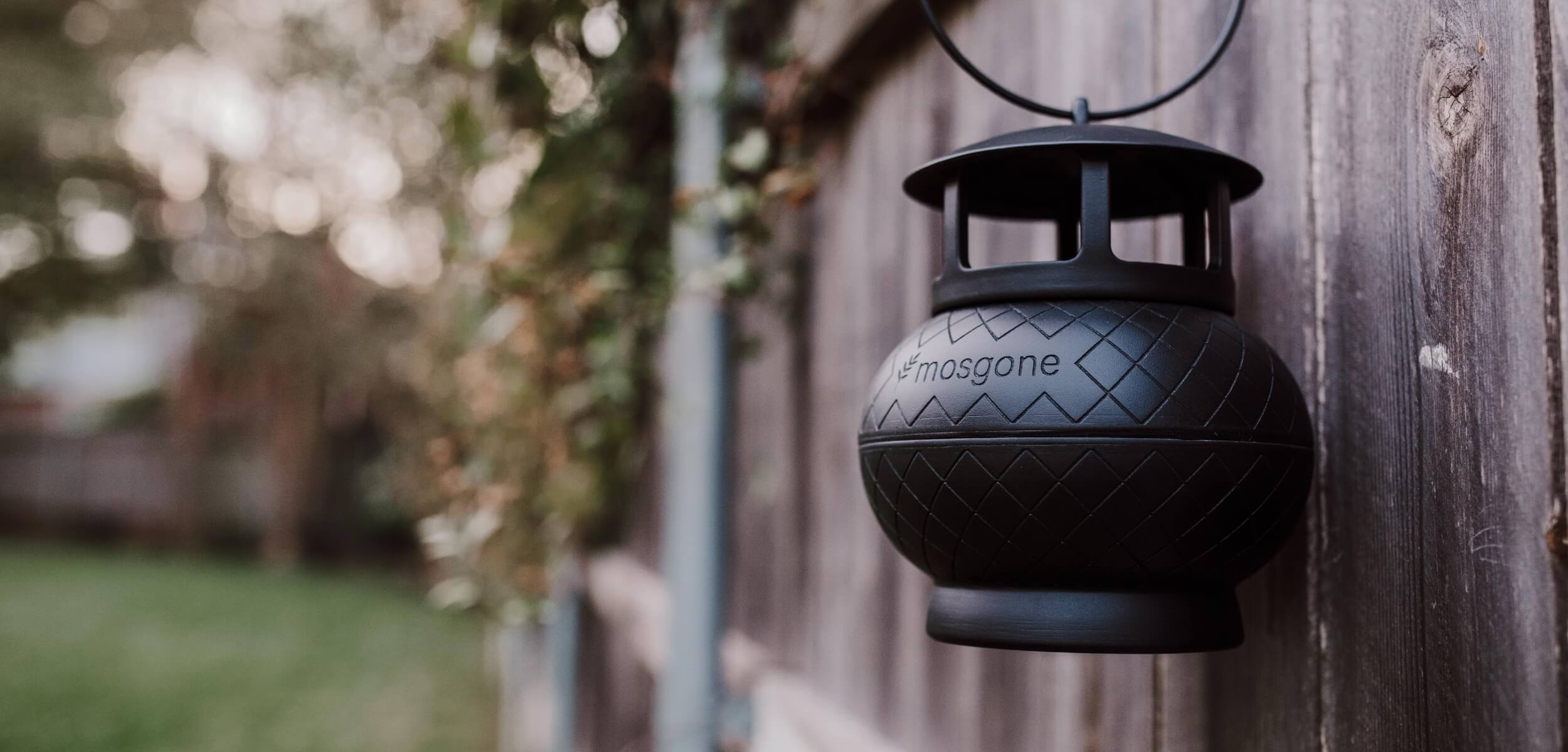 A black, round Mosgone lantern hangs on a wooden fence, with blurred greenery and a yard visible in the background. The lantern features a geometric pattern and is in sharp focus.