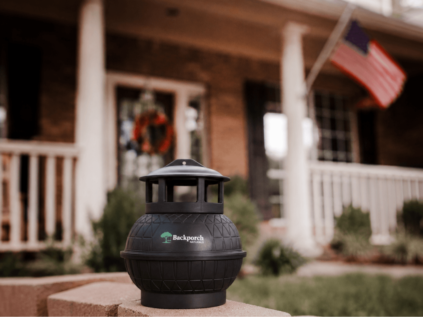 A black Backporch Naturals Mosquito Starter Kit by Mosgone Mosquito Control Products sits on a porch ledge, with a brick house, white railing, and an American flag blurred in the background.