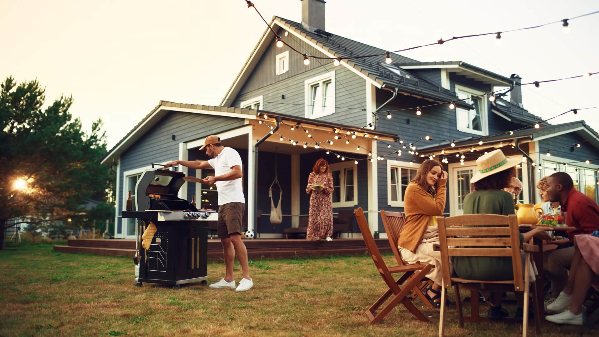 A group of people enjoying a backyard barbecue. One person grills food, while others sit at a wooden table, talking and smiling. String lights hang above, and a house with a porch and garden is in the background.