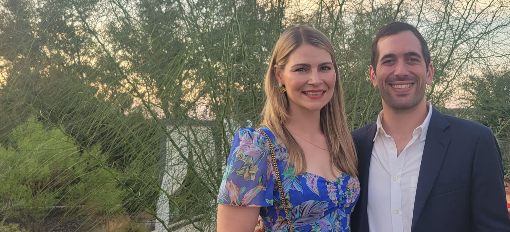 A smiling couple poses outdoors in front of green foliage. The woman has long blonde hair and wears a blue floral dress; the man has short dark hair and wears a navy blazer over a white shirt.